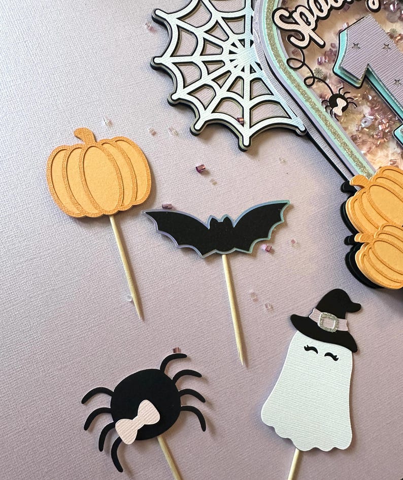 May include: Five Halloween-themed cake toppers. The toppers are made of paper and feature a pumpkin, a bat, a spider, a ghost wearing a witch's hat, and a spider web.