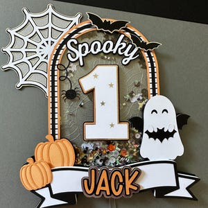 May include: A spooky Halloween cake topper with a white number one, a black spider, a white ghost with a bat, and a banner that says "Spooky Jack".