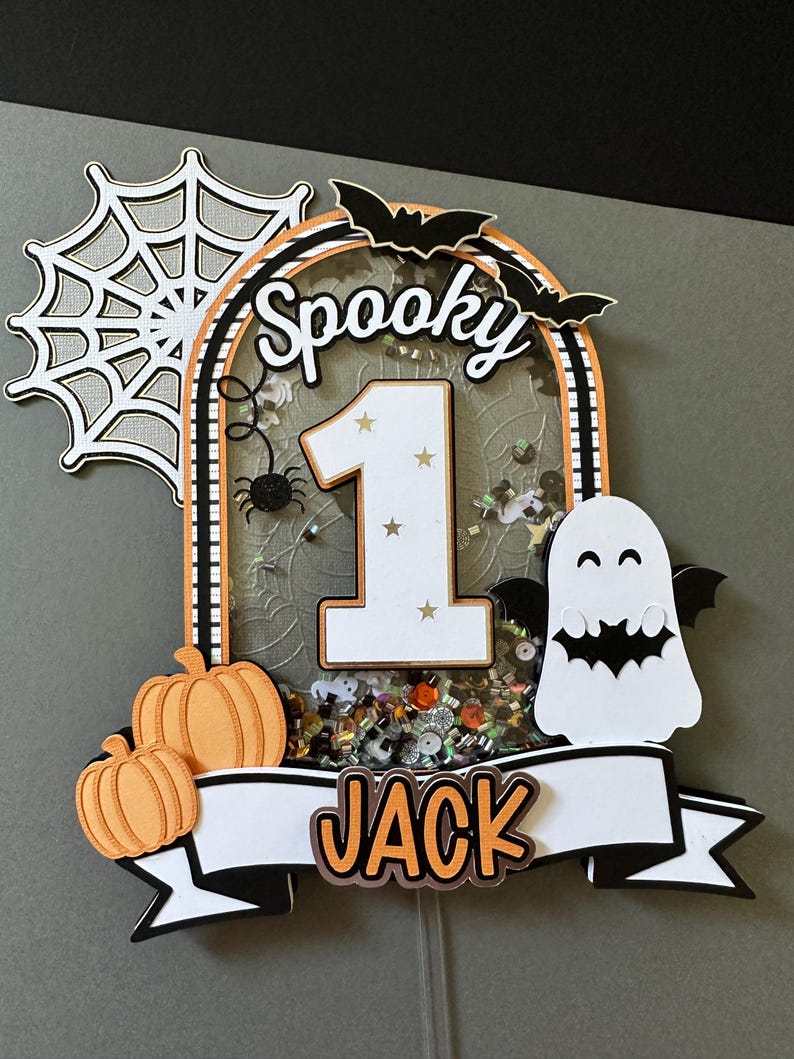 May include: A spooky Halloween cake topper with a white number one, a black spider, a white ghost with a bat, and a banner that says "Spooky Jack".