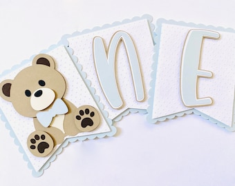 May include: A blue and white banner with the word "one" spelled out in blue letters. The banner features a brown teddy bear with a blue bow tie and brown paw prints.