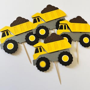 May include: Five yellow and gray dump truck cupcake toppers with brown cargo. The trucks have black wheels with yellow centers. Each topper is attached to a wooden toothpick.