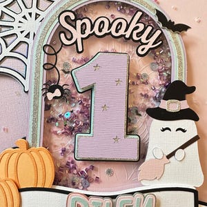 May include: A pink and green cake topper with a glittery background, a spider, a ghost, a pumpkin, and the words "Spooky" and "Riley".