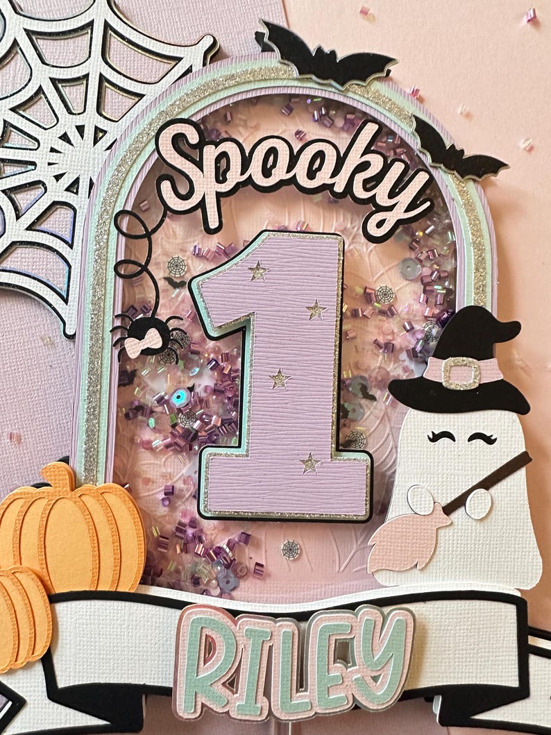 May include: A pink and green cake topper with a glittery background, a spider, a ghost, a pumpkin, and the words "Spooky" and "Riley".