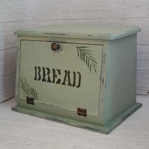 Könnte beinhalten: Ein salbeigrüner Brotkasten aus Holz mit einer nach unten aufklappbaren Fronttür, auf der das Wort BREAD in dunkler Schrift steht. Der Kasten hat ein abgenutztes Finish, zwei Metallscharniere unten und einen runden Knopf oben mittig.