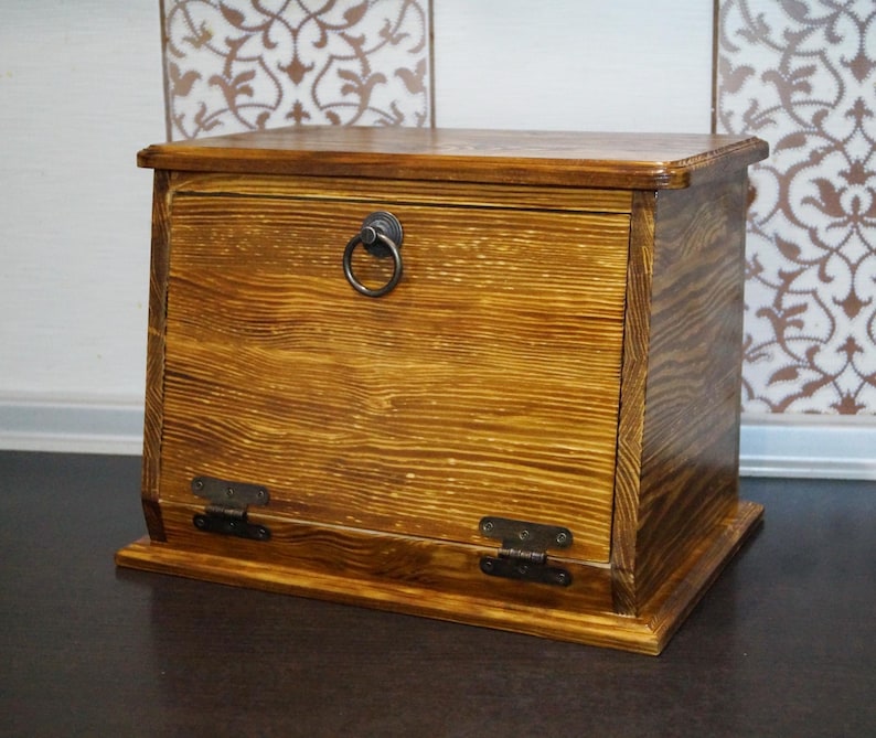Puede incluir: Una caja de almacenamiento de madera con tapa con bisagras y un asa de anillo met&aacute;lico. La caja est&aacute; te&ntilde;ida en un color marr&oacute;n dorado c&aacute;lido, con vetas de madera visibles. Cuenta con bisagras met&aacute;licas y una forma rectangular.