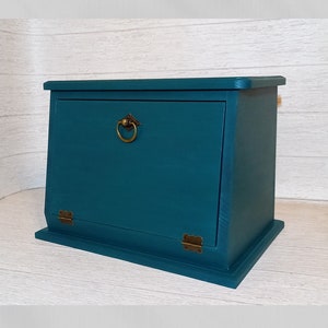 May include: Teal wooden storage box with a hinged door and brass fittings. The box has a rectangular base and a sloped top. The door features a brass ring pull. The box is painted in a solid teal colour.