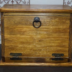 Puede incluir: Una caja de madera con tapa con bisagras y tirador de anillo. La caja es de madera marr&oacute;n claro con veta visible y est&aacute; te&ntilde;ida con un tono c&aacute;lido y dorado. Tiene bisagras met&aacute;licas y un tirador de anillo met&aacute;lico.