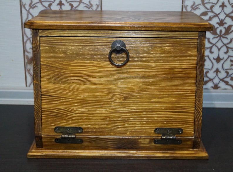 Puede incluir: Una caja de madera con tapa con bisagras y tirador de anillo. La caja es de madera marr&oacute;n claro con veta visible y est&aacute; te&ntilde;ida con un tono c&aacute;lido y dorado. Tiene bisagras met&aacute;licas y un tirador de anillo met&aacute;lico.