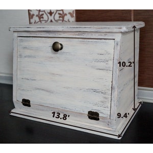 Puede incluir: Una panera de madera pintada de blanco con un acabado envejecido. La caja tiene una puerta con bisagras, un pomo de metal y mide 35 cm de ancho, 24 cm de profundidad y 26 cm de alto.