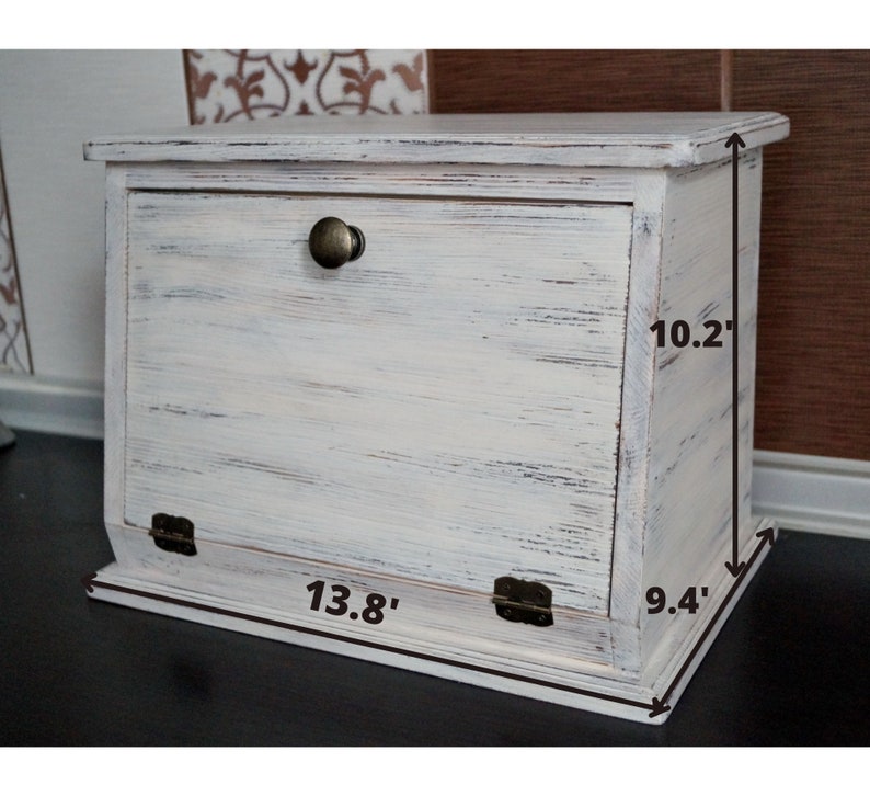 Puede incluir: Una panera de madera pintada de blanco con un acabado envejecido. La caja tiene una puerta con bisagras, un pomo de metal y mide 35 cm de ancho, 24 cm de profundidad y 26 cm de alto.