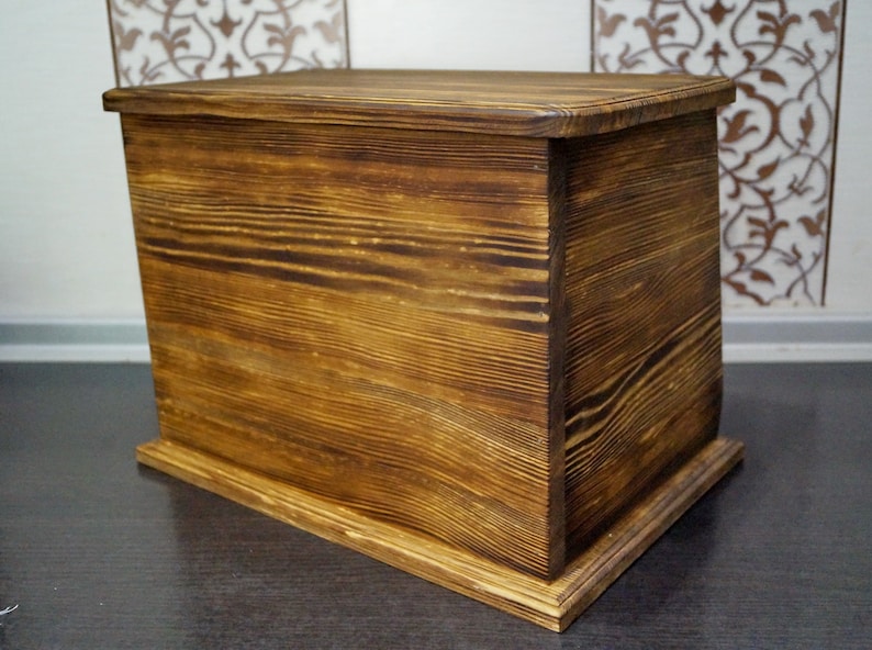 Puede incluir: Un cofre de madera rectangular con un acabado marr&oacute;n dorado c&aacute;lido. El cofre tiene una tapa y se apoya sobre una base ligeramente elevada. La veta de la madera es visible, agregando textura. El cofre probablemente se usa para almacenamiento.