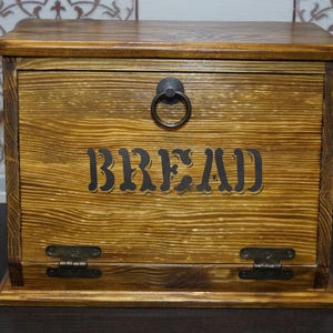 Puede incluir: Caja de pan de madera con tapa con bisagras y asa de anillo met&aacute;lico. La parte frontal de la caja presenta la palabra "BREAD" en una fuente oscura y estilizada. La caja tiene un c&aacute;lido acabado de veta de madera natural.