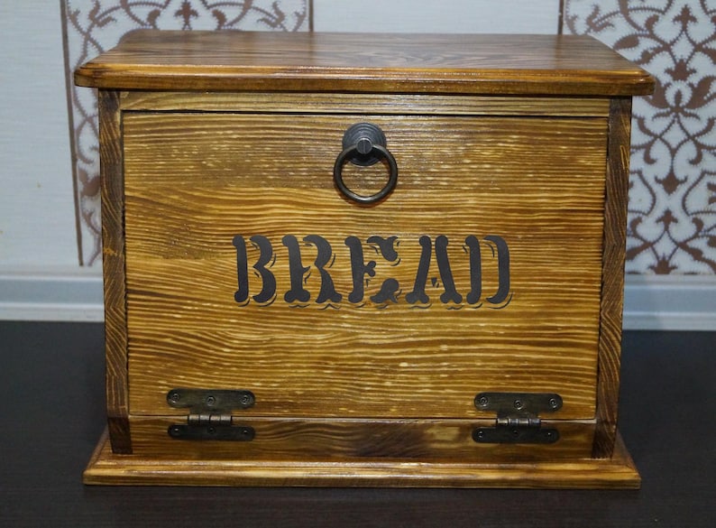 Puede incluir: Caja de pan de madera con tapa con bisagras y asa de anillo met&aacute;lico. La parte frontal de la caja presenta la palabra "BREAD" en una fuente oscura y estilizada. La caja tiene un c&aacute;lido acabado de veta de madera natural.