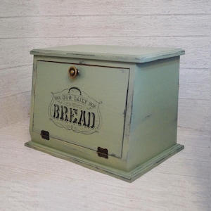 May include: A vintage-style bread box painted in a soft green hue with distressed edges. The front door features the words "OUR DAILY BREAD" in a decorative black font. It has a brass knob and metal hinges.