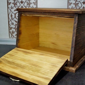 Puede incluir: Una panera r&uacute;stica de madera con tapa abatible, hecha de madera de color claro con un marco te&ntilde;ido oscuro. La caja est&aacute; abierta, revelando el interior. Se puede ver la veta de la madera.