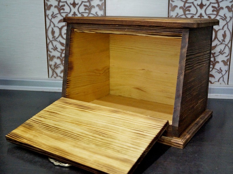 Puede incluir: Una panera r&uacute;stica de madera con tapa abatible, hecha de madera de color claro con un marco te&ntilde;ido oscuro. La caja est&aacute; abierta, revelando el interior. Se puede ver la veta de la madera.