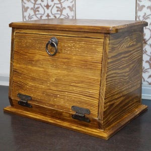 Puede incluir: Una panera de madera con una puerta frontal con bisagras y un tirador de anillo. La panera est&aacute; hecha de madera marr&oacute;n claro con vetas visibles y detalles en marr&oacute;n oscuro. Las bisagras y el tirador son de metal oscuro.