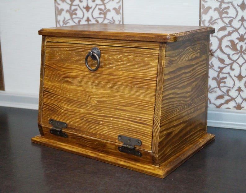Puede incluir: Una panera de madera con una puerta frontal con bisagras y un tirador de anillo. La panera est&aacute; hecha de madera marr&oacute;n claro con vetas visibles y detalles en marr&oacute;n oscuro. Las bisagras y el tirador son de metal oscuro.