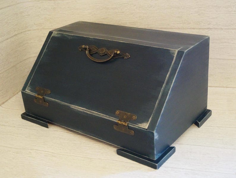 Puede incluir: Una caja de madera azul marino con tapa inclinada, bisagras de lat&oacute;n y un asa decorativa. La caja tiene un acabado envejecido, lo que le da un aspecto vintage. La caja es rectangular y se apoya sobre cuatro peque&ntilde;os pies.