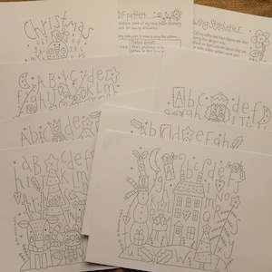 Pode incluir: Uma coleção de padrões de bordado desenhados à mão em cartões brancos. Os desenhos incluem alfabetos com tema natalino, elementos decorativos e a palavra "Christmas". Outros cartões apresentam alfabetos com ilustrações caprichosas. O texto "Wing Stitcheries" também é visível.