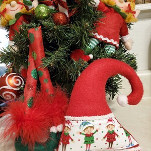 May include: Three red and green felt elf ornaments with white trim. One elf is wearing a red hat with white trim and has a white pom-pom on the top. The other two elves are wearing green hats with white trim and have red and white striped candy canes. The elves are all sitting on a green Christmas tree.