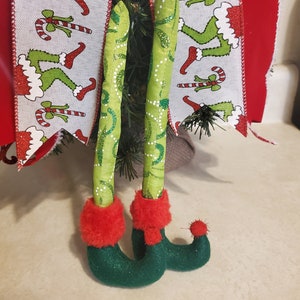 May include: A pair of green elf legs with red fuzzy boots. The legs are made of fabric and have a sparkly finish. The boots have a pointed toe and a small red pom-pom on the top.
