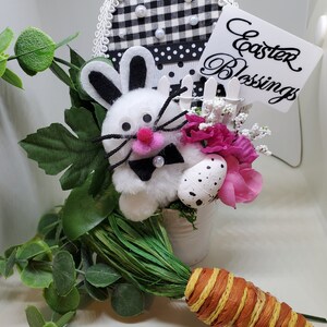 May include: White bunny with black ears and a pink nose in a white bucket with pink flowers and a white picket fence. The bucket is sitting on a white surface with a carrot and green foliage. A sign reads "Easter Blessings".