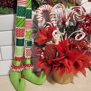 May include: A pair of green elf legs with red and white striped fabric and green and white patterned fabric. The legs are standing next to a red poinsettia centerpiece with white candy cane swirls and red berries.