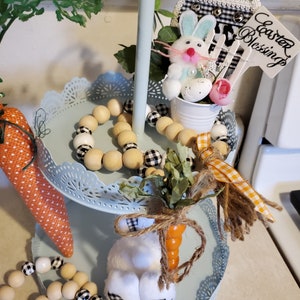 May include: A three-tiered stand with a light blue finish and white lace trim. The top tier features a white bunny figurine, a small white pot with a green plant, and a sign that reads "Easter Blessings". The middle tier has a wooden bead garland with black and white checkered beads. The bottom tier has a carrot-shaped wooden dowel with a white bunny figurine and a wooden bead garland with black and white checkered beads.