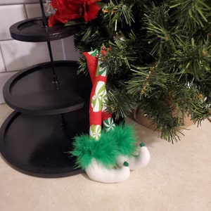 Könnte beinhalten: Ein Paar Elfenbeine mit grün-weiß gestreiften Strümpfen und grünen, flauschigen Stiefeln. Die Beine hängen an einem Weihnachtsbaum.