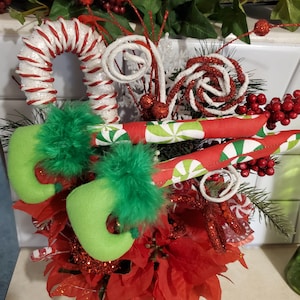 May include: A festive Christmas arrangement featuring candy canes, red and white swirled lollipops, and elf shoes. The arrangement includes red poinsettias, berries, and green foliage, creating a cheerful holiday display.