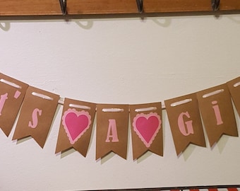 May include: A brown paper banner with pink lettering that reads "It's a Girl" with pink baby bottle icons on each end.