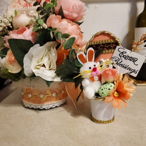 May include: A white bunny rabbit with orange ears sits in a white pot with a gold trim. The pot is filled with pink, orange, and green flowers. The bunny is holding a green speckled egg. The pot is sitting on a table next to a larger pot with pink and white flowers. The larger pot has a peach colored ribbon with gold trim and a white lace trim. The text "Easter Blessings" is written on a white tag.