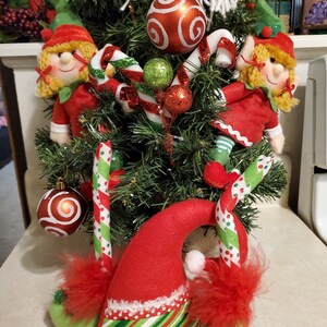 Könnte beinhalten: Ein Weihnachtsbaum-Mittelstück mit zwei Elfen, Zuckerstangen und roten und grünen Kugeln. Die Elfen tragen rote und weiße Mützen und haben blondes Haar. Der Baum ist mit roten und weißen Zuckerstangen und roten und grünen Kugeln geschmückt.