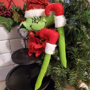 Könnte beinhalten: Zwei grüne Filz-Weihnachtsornamente im Grinch-Stil in Form von Zuckerstangen mit roten und weißen Weihnachtsmannmützen. Die Ornamente werden auf einem schwarzen, mehrstufigen Ständer präsentiert.