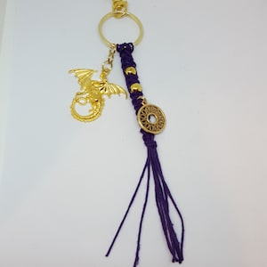 May include: A gold dragon keychain with a purple tassel and a gold circle charm. The keychain is attached to a gold split ring.
