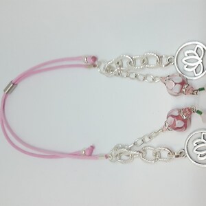 Könnte beinhalten: Eine rosa Schnurkette mit zwei silbernen Anhängern mit Lotusblumen-Design. Die Anhänger sind an einer silbernen Kette mit rosa und weißen Glasperlen befestigt.