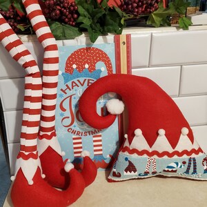 May include: Two red and white striped elf legs with red felt shoes and a red felt elf hat with a white trim and a blue and white patterned base. The hat has the text "Have a Joyful Christmas" on a blue background.