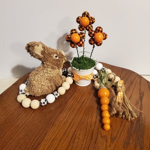 May include: A brown bunny rabbit figurine sits on a wooden surface with a garland of white and black checked beads and wooden beads. A small white bucket with a green moss base and three orange and black checked flowers sits on the surface. A carrot-shaped garland with orange beads hangs from the bucket.