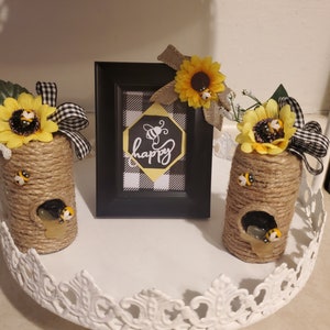Puede incluir: Dos tarros decorativos envueltos en cuerda y coronados con girasoles y abejas. Los tarros están en una bandeja blanca con un marco a cuadros blanco y negro que dice "happy" con una abeja dentro de un hexágono.