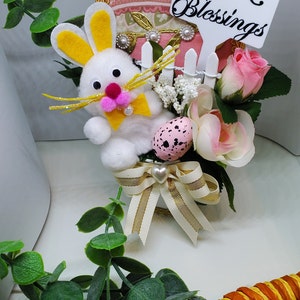 Op de afbeelding: Een wit konijn met gele oren en een roze strik zit in een witte schutting-plantenbak. De plantenbak is versierd met een gouden lint en een hartvormige hanger. De plantenbak is omringd door roze en witte bloemen. Een gevlekt ei zit in de plantenbak. Een bordje staat "Easter Blessings".
