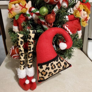 May include: A Christmas tree decoration featuring two red and white elf figures with blonde hair, a red and white candy cane, and a red and white ornament. The elf figures are wearing leopard print pants and boots.