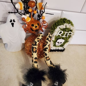 May include: Halloween decoration featuring a witch with leopard print leggings and black boots with feather trim. The witch is holding a pumpkin with a jack-o-lantern face and a twig with orange and white candy corn. There is also a ghost and a green tombstone with the text "RIP" and a skull.