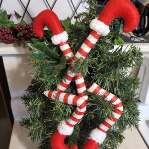 Könnte beinhalten: Zwei rote und weiße Zuckerstangen-Elfenbeine, die um einen kleinen künstlichen Weihnachtsbaum gewickelt sind. Die Elfenbeine sind aus Filz und haben einen weißen Pelzbesatz.