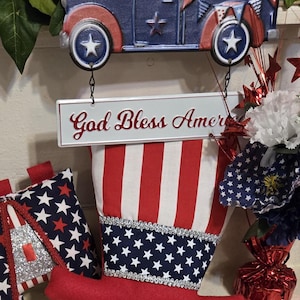 Puede incluir: Un colgador de puerta de temática patriótica con un camión rojo, blanco y azul, un letrero que dice "God Bless America" y una media roja, blanca y azul.