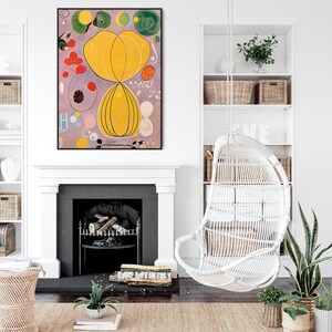 May include: A framed abstract painting featuring a central yellow circular shape, surrounded by smaller circles and other shapes in shades of pink, green, and blue. The painting is displayed against a white wall, with a white fireplace and a white hanging chair in the background.