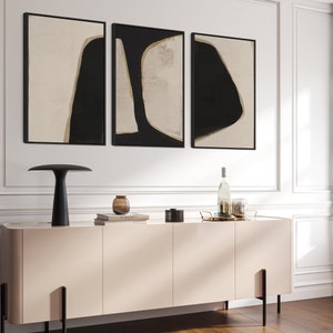 May include: Three abstract art prints featuring black and beige shapes on a white background. The prints are framed in black and hung on a white wall above a light beige cabinet.