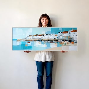 May include: A wide rectangular framed painting depicting a coastal village scene with white buildings featuring orange roofs, small boats floating on calm blue water, and abstract geometric shapes in shades of light blue, white, and tan.