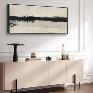 May include: Abstract artwork in a black frame with a thick black band on a cream background. A black lamp, a vase, books, and a cabinet with black legs are in the foreground. The cabinet is approximately 150 cm long.