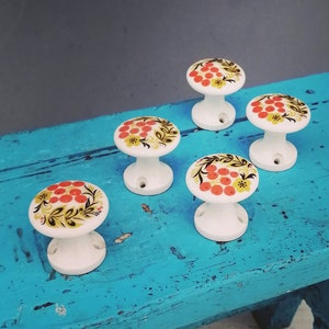 Puede incluir: Seis pomos de cerámica blanca con un diseño floral en tonos rojos, amarillos y verdes. Los pomos están dispuestos en semicírculo sobre un taburete de madera pintado de azul.